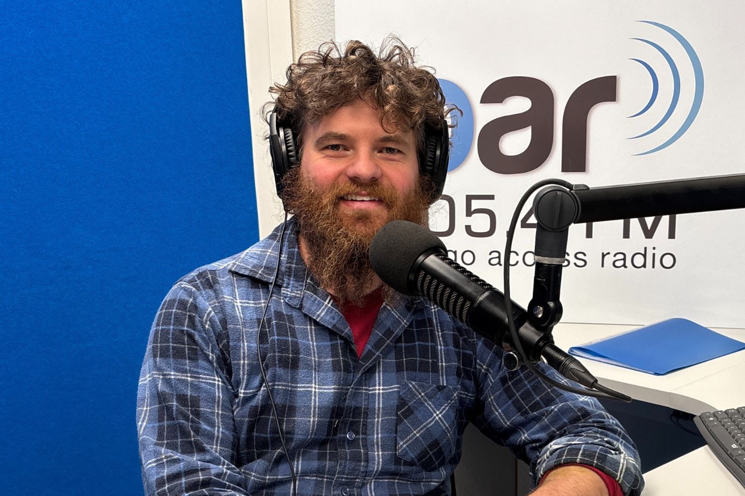 Photo of Kieran in the OAR FM Studio in front of an OAR FM Sign and Microphone