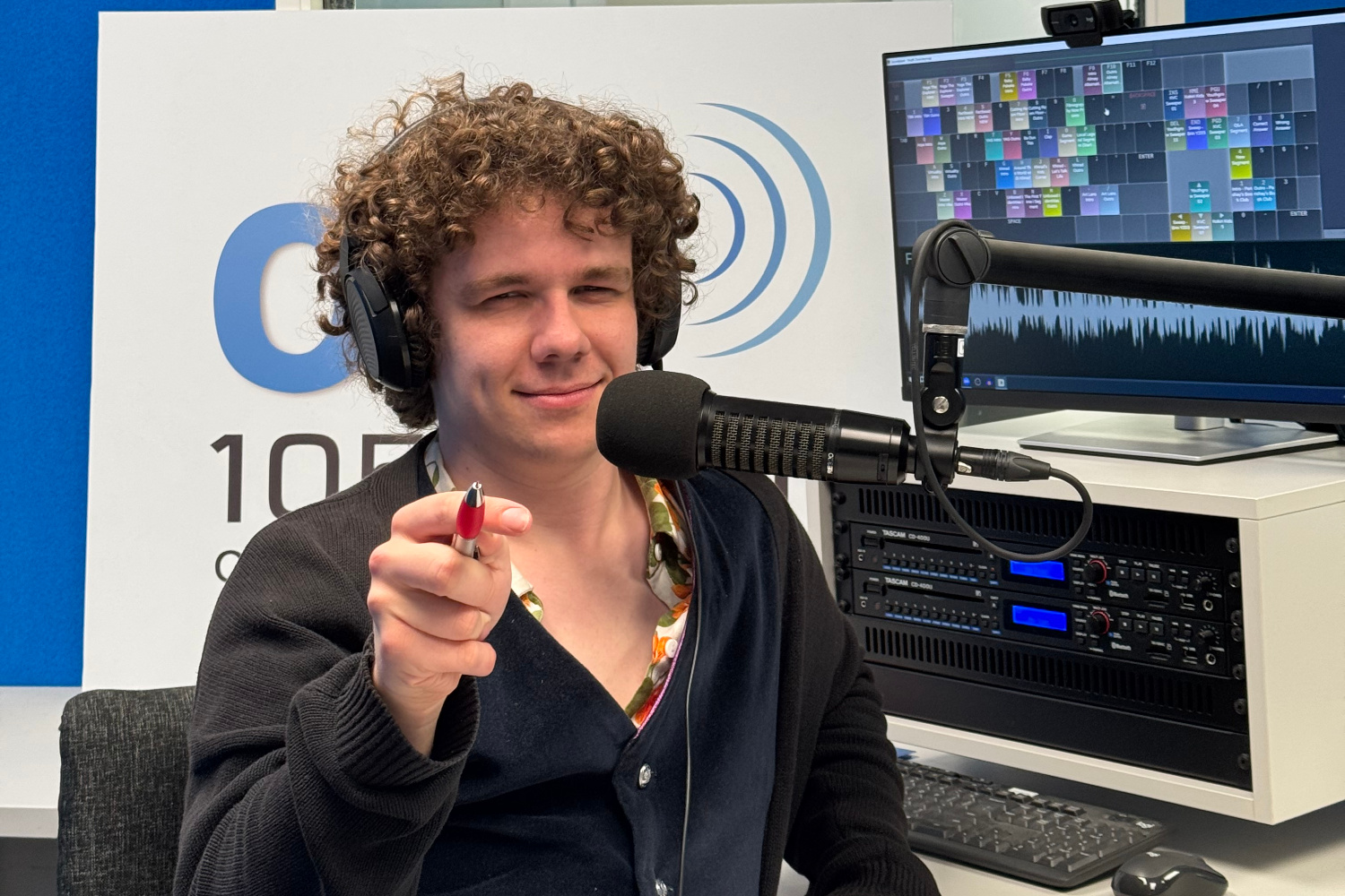 A photo of Harry Almey in OAR FM Studio in front of a microphone and pointing a pen at the camera